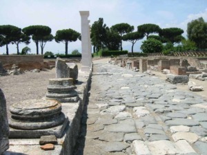 Antico sito archeologico di Roma antica con pavimentazione in pietra e resti di colonne, immerso nel verde di pino marittimo e cipressi, esempio di archeologia e storia romana.