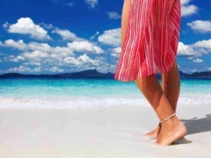 Passeggiata sulla spiaggia con abito estivo leggero e ciambella alla caviglia, ambiente marino con acque cristalline e cielo nuvoloso, ideale per relax e vacanze estive.