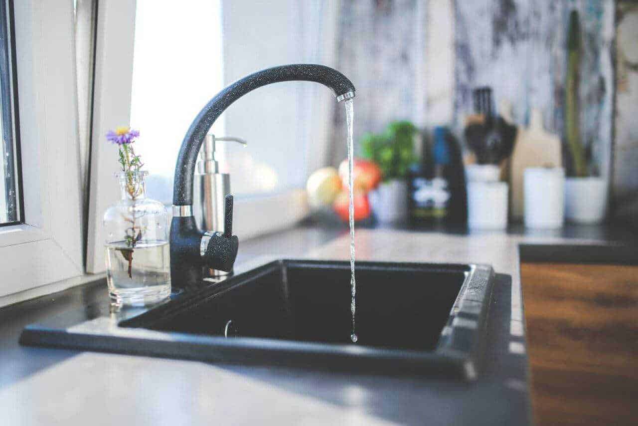 Lavandino da cucina con rubinetto aperto e acqua che scorre, pianta fiorita in bottiglia di vetro, sfondo con utensili e piante, ambiente domestico luminoso e accogliente.