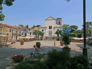 Piazza centrale di un piccolo paese italiano con chiesa storica, caffè all'aperto e persone che passeggiano, atmosfera tranquilla e pittoresca, perfetta per scoprire la cultura locale.