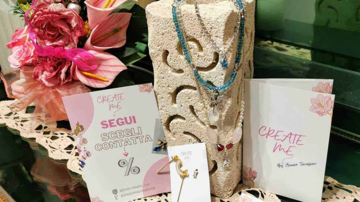 Lucine e collane di bijoux colorati esposte su un supporto di pietra, con fiori rosa e biglietti promozionali, creano un'ambientazione elegante per il negozio di accessori moda.