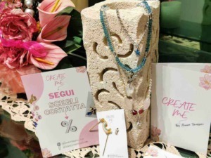 Lucine e collane di bijoux colorati esposte su un supporto di pietra, con fiori rosa e biglietti promozionali, creano un'ambientazione elegante per il negozio di accessori moda.
