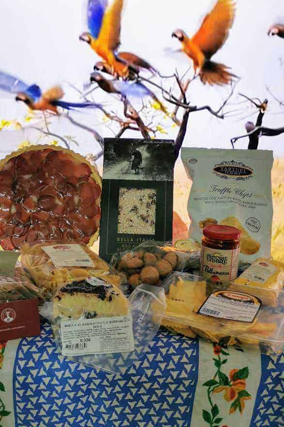Mater Umbria vendita prodotti tipici italiani 2 Prodotti tipici italiani online: Voli di uccelli colorati che si librano nel cielo sopra un'ampia selezione di prodotti alimentari italiani, tra cui dolci, biscotti e conserve, con uno sfondo di cielo e natura.