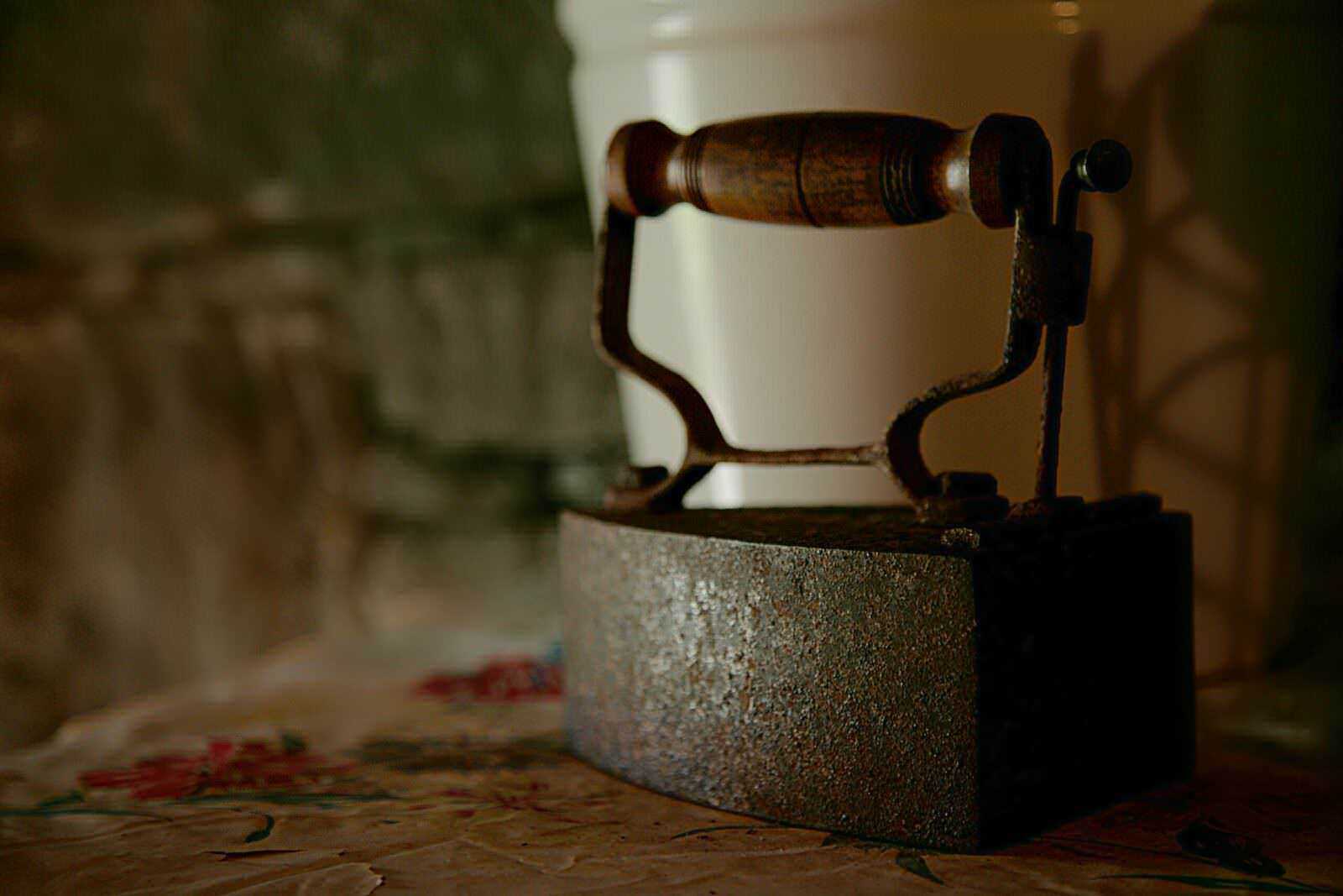 Più dettagliato: Antica piastra a vapore in ferro battuto con manico in legno, posizionata su una superficie di piastrelle decorative, ambiente rustico e vintage, perfetta per decorazioni di antiquariato o collezionismo.