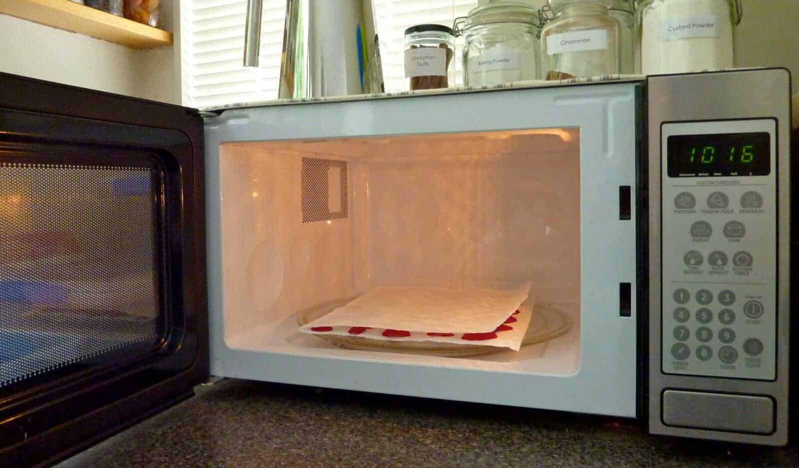 Come conservare i fiori recisi, 7 metodi facili 3 L'immagine mostra un forno a microonde con un piatto di carta e un tovagliolo all'interno, pronto per essere riscaldato o cotto. Sul ripiano superiore si vedono barattoli di spezie e aromi, come cannella e anice, in contenitori di vetro trasparente. La scena rappresenta un ambiente domestico e cucina, perfetta per contenuti legati a ricette, cucina casalinga e tecniche di riscaldamento alimentare.