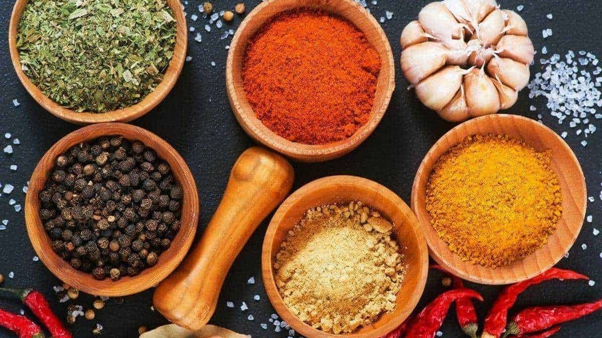 Miscela di spezie e aromi in ciotole di legno con aglio, pepe nero, curcuma, paprika e cumino, su sfondo nero.