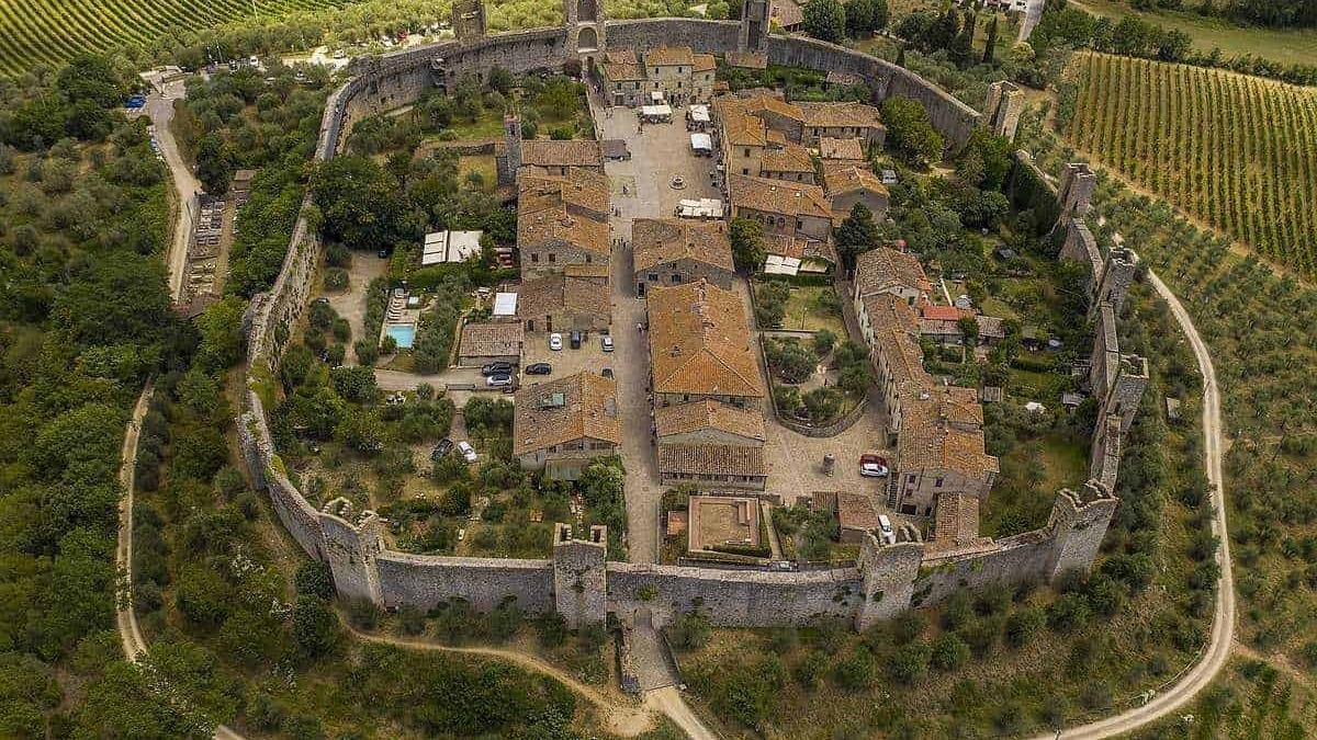 Antica fortezza medievale circondata da mura in pietra, situata tra vigneti e colline verdi in Italia, con case e cortili all’interno delle mura.
