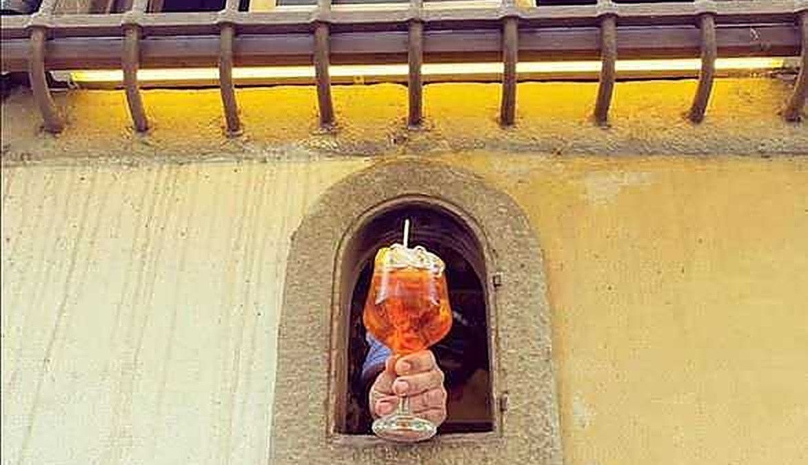 Aperitivo in un pub, mano che tiene un drink color ambra in un bicchiere con cannuccia, vista da una finestra su una strada cittadina, atmosfera accogliente e convivialità.