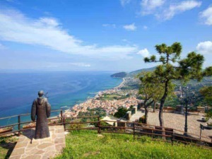 Vista panoramica di una costa con mare e città, con statue e panchine lungo il percorso, su un rilievo con alberi e uno sfondo di cielo blu e nuvole.