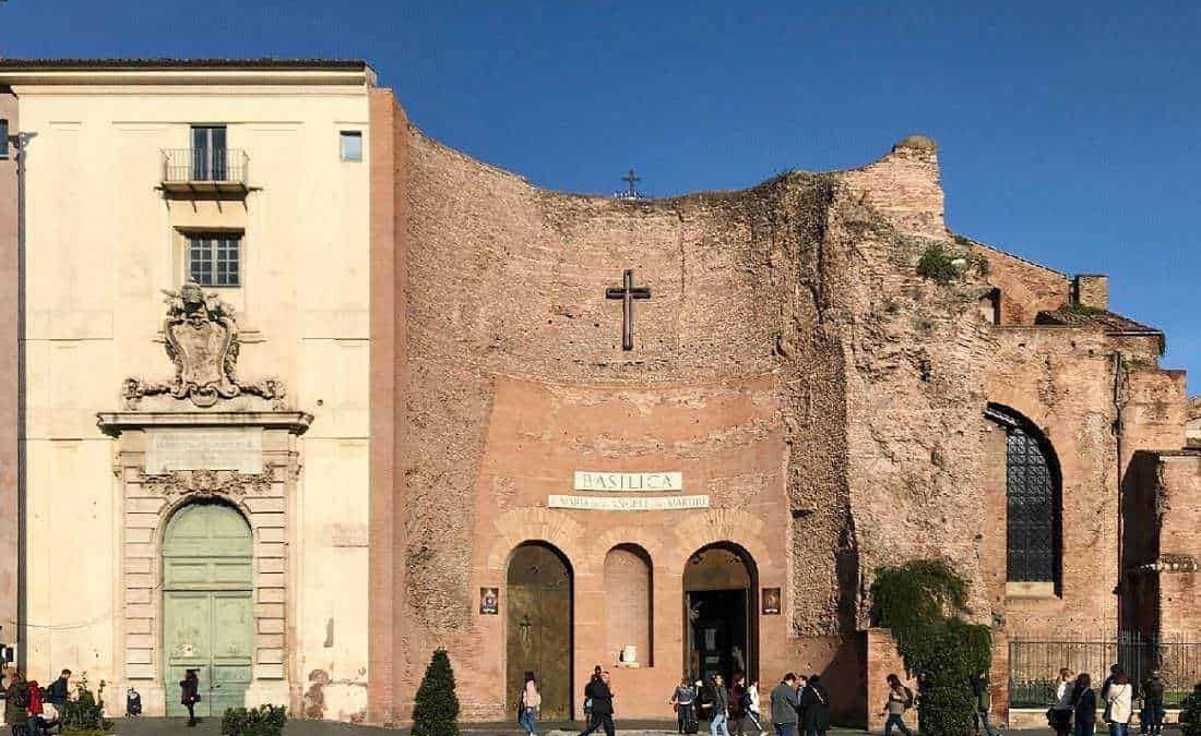 Antica chiesa e rovine storiche di Roma, ingresso alla basilica con archi e croce, scena urbana con persone, cielo blu e elementi di architettura religiosa, centro storico, monumento storico.