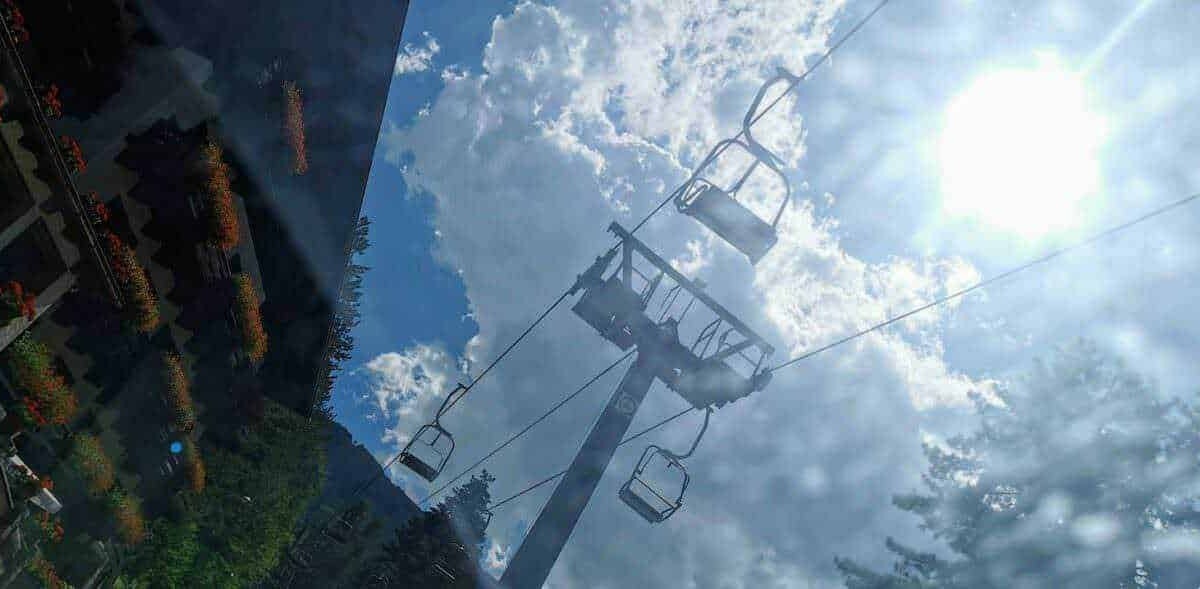 Cavo funiviale in montagna con sedie per sci e outdoor, cielo nuvoloso e sole splendente, ambiente alpino con alberi e edifici circostanti.