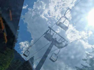 Cavo funiviale in montagna con sedie per sci e outdoor, cielo nuvoloso e sole splendente, ambiente alpino con alberi e edifici circostanti.