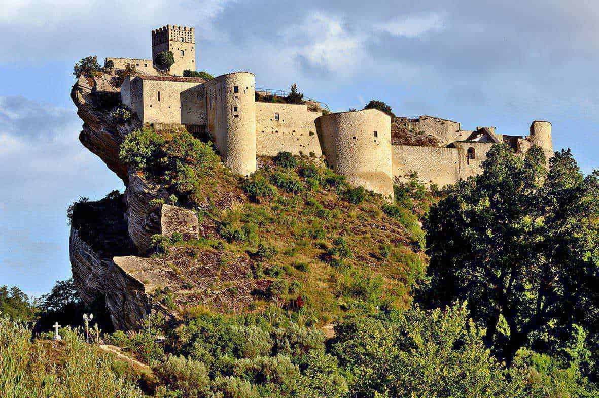 Dove andare in Abruzzo montagna estate 3 Castello medievale su scogliera con vegetazione circostante e cielo nuvoloso, esempio di architettura storica italiana e attrazione turistica.