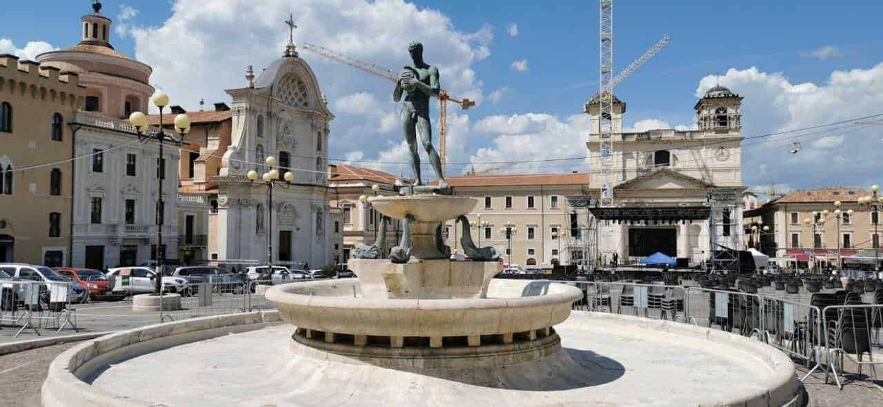Dove andare in Abruzzo montagna estate 7 Fonte Piazza con fontana e statue al centro, scena urbana di Cagliari con edifici storici e palco per eventi, cielo sereno e traffico, rappresentativa del cuore della città.