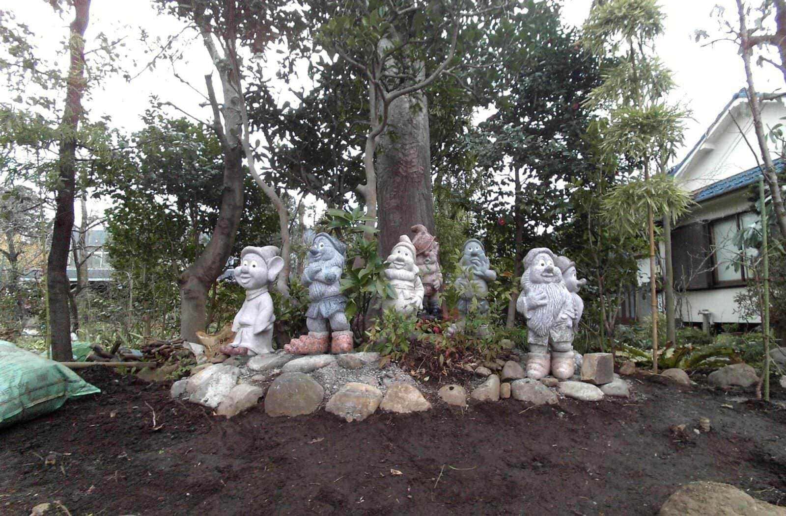 Origine Nani da giardino da antica Roma a oggi! 4 Colorata statua di sette nani della fiaba di Biancaneve in giardino, circondata da piante e alberi, con una casa sullo sfondo, creando un’atmosfera fiabesca e naturale.