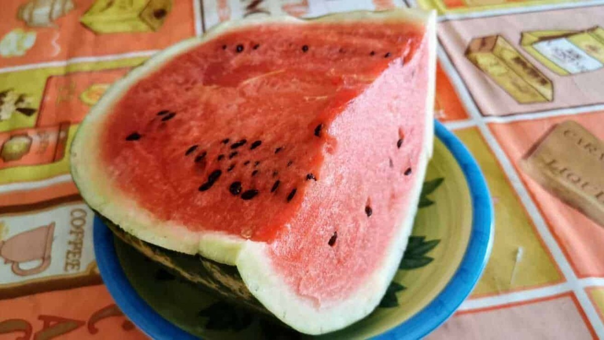 Succosa fetta di anguria fresca e succulenta su piatto blu, con sfondo di tovaglia colorata a tema frutta e bevande, perfetta per l'estate.