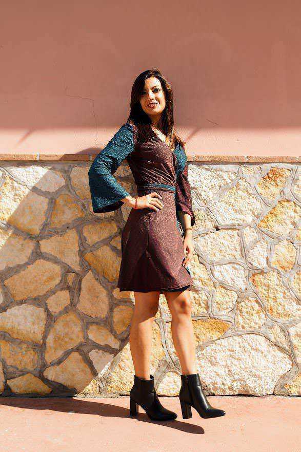 Elegante donna che indossa un vestito elegante e stivali neri, posando con sicurezza davanti a un muro di pietra e una parete rosa, perfetta per contenuti di moda e stile di vita.