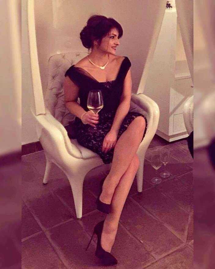 Elegante donna in abito nero con calze a rete e tacchi a spillo, seduta su una poltrona con calice di vino, atmosfera raffinata e glamour.