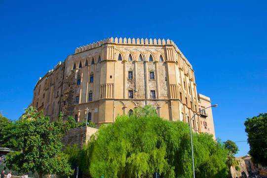 25 luoghi da visitare a Palermo e dintorni in stile arabo normanno 4 Antico castello Medievale con mura imponenti e torri, circondato da alberi verdi e cielo azzurro, sito storico in Italia, esempio di architettura e cultura medievale.