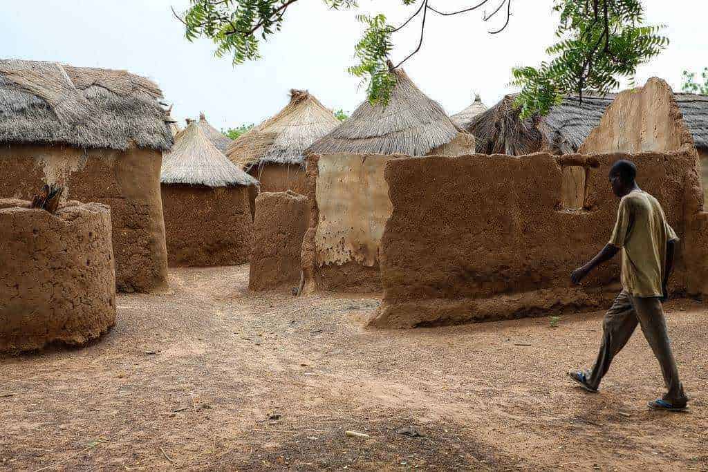 Paesaggi agricoli tropicali e subtropicali: Vecchi alloggi tradizionali in terra e paglia nel villaggio africano, con capanne dal tetto di paglia e strutture di argilla, immerso nella natura, con un uomo che attraversa la strada sterrata.