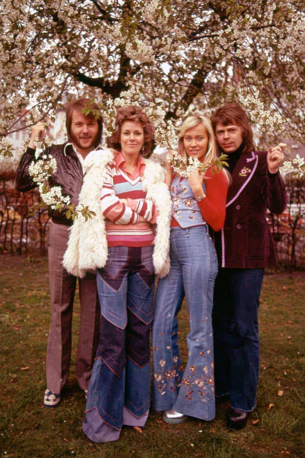 Immagine di un gruppo di amici o famiglia che si gode una giornata primaverile con alberi in fiore, in un parco. Atmosfera anni '70, abbigliamento colorato e stile vintage, perfetto per articoli su moda retrò e lifestyle.