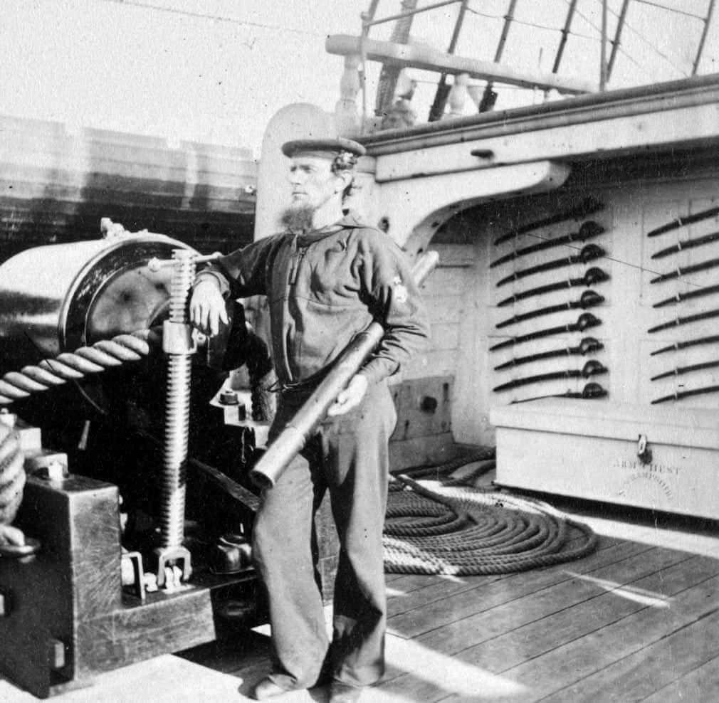 Antico uomo in uniforme a bordo di una nave storica, vicino a equipaggiamento navale e cannoni, scenario marittimo vintage.