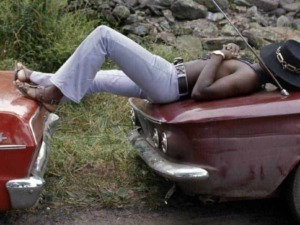 Auto d'epoca e donna sdraiata con cappello, scena rilassante in natura, fotografia vintage di auto e moda, perfetta per appassionati di automobili classiche e stile retrò.