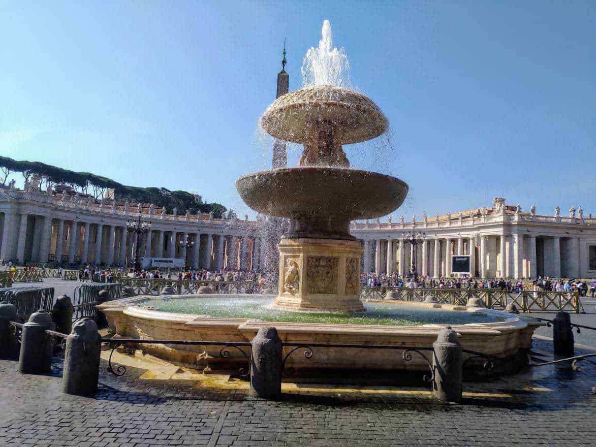 Fontana di Piazza San Pietro a Vatican City, simbolo di Roma, con statua decorata e acqua che spruzza verso il cielo, circondata da edifici storici e visitatori, esempio di arte e architettura classica.