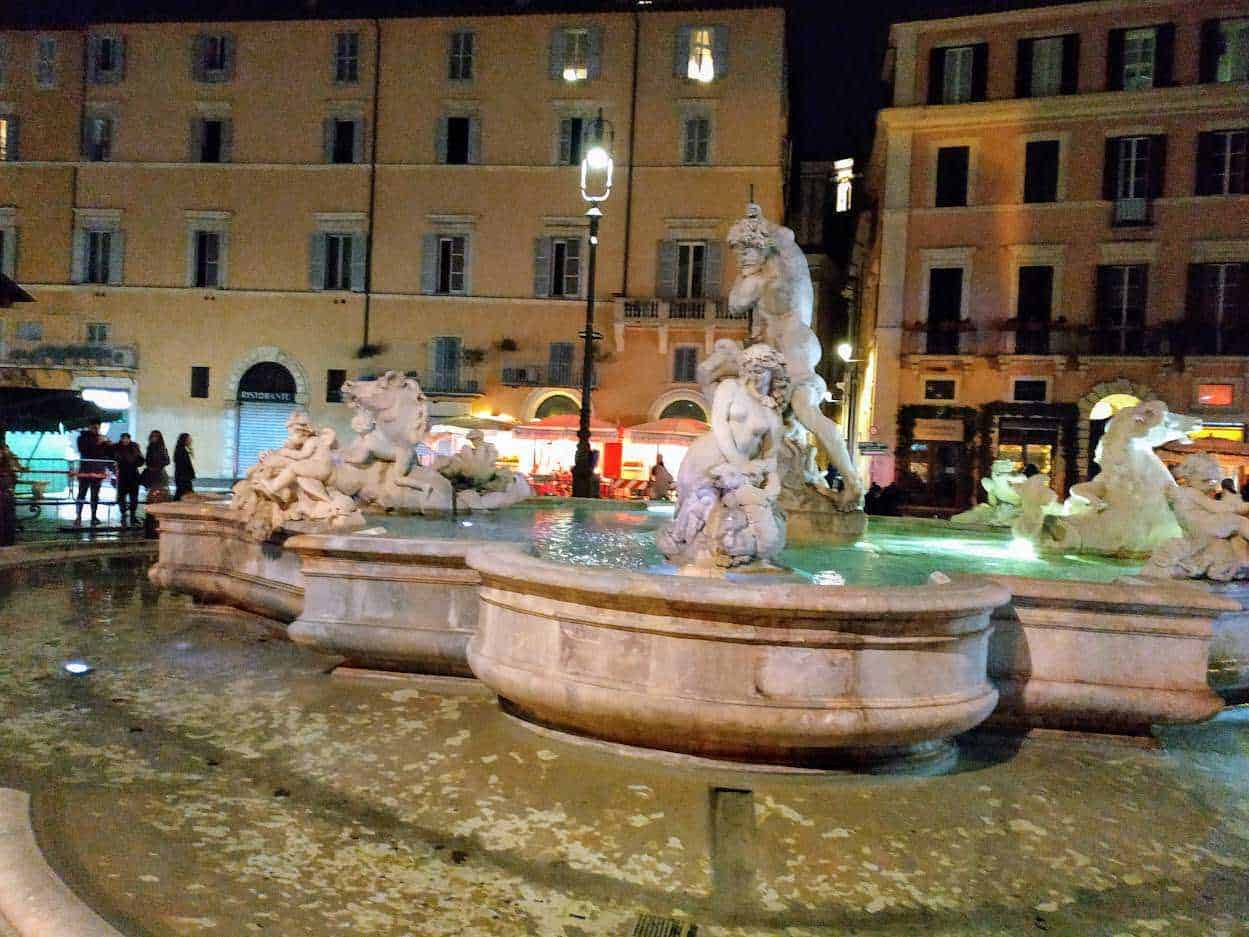 Fountain with statues in Piazza Navona, Rome at night illuminated by streetlights, lively atmosphere with tourists and locals, baroque architecture background.