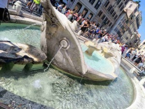 Fontana storica nel centro di Roma con persone che la ammirano e si rilassano, in un contesto urbano affollato, con edifici storici sullo sfondo.