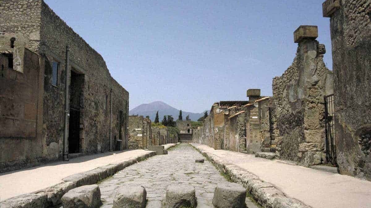 Antica strada romana tra macerie e rovine di Pompei, vulcano in lontananza, sito archeologico storico italiano.