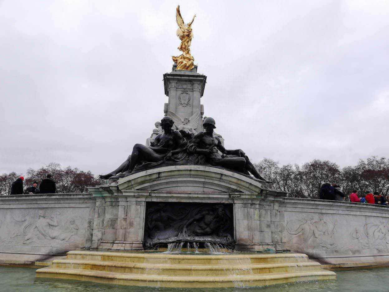 Statua monumentale con figure in bronzo e dettagli dorati, scenario di piazza pubblica con persone che visitano il monumento, ambientazione storica e culturale