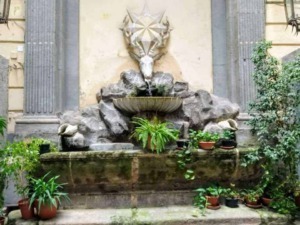 Fontana decorativa con elementi di pietra, dettagli artistici e piante in vaso, in un cortile storico, ideale per foto di design di giardini e architettura urbana.