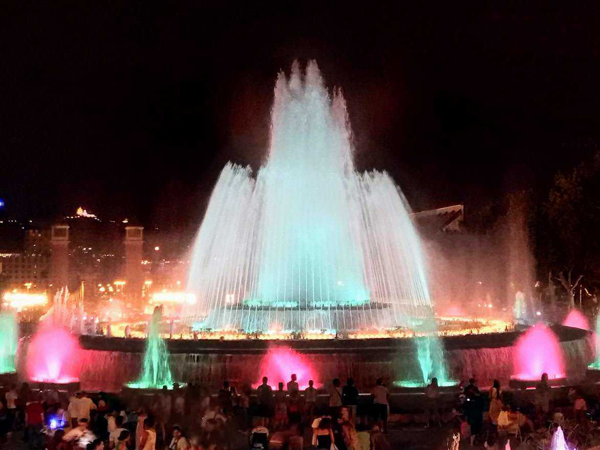 Fontana luminosa di notte con giochi d'acqua colorati e pubblico che ammira lo spettacolo, illuminata da luci multicolori, ambientata in un'area urbana di sera.