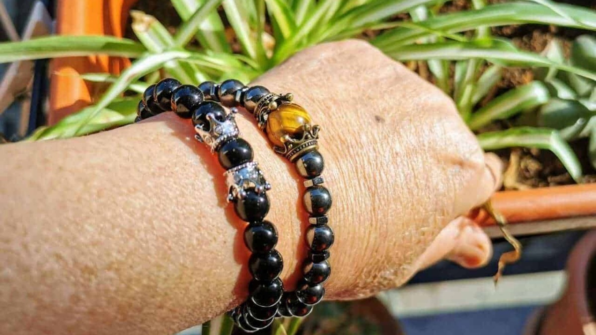 Braccialetti di perle nere con dettagli oro e argento, posizionati su un polso di pelle, con piante verdi sullo sfondo, stile elegante e di moda.