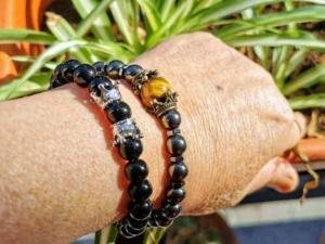 Braccialetti di perle nere con dettagli oro e argento, posizionati su un polso di pelle, con piante verdi sullo sfondo, stile elegante e di moda.