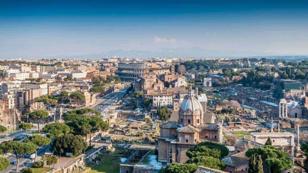 Roma dall'alto con il Colosseo e i resti storici al centro, scenario urbano ricco di storia e cultura, con monumenti antichi e moderni, sotto un cielo azzurro e luminoso.