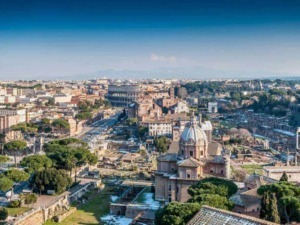 Roma dall'alto con il Colosseo e i resti storici al centro, scenario urbano ricco di storia e cultura, con monumenti antichi e moderni, sotto un cielo azzurro e luminoso.