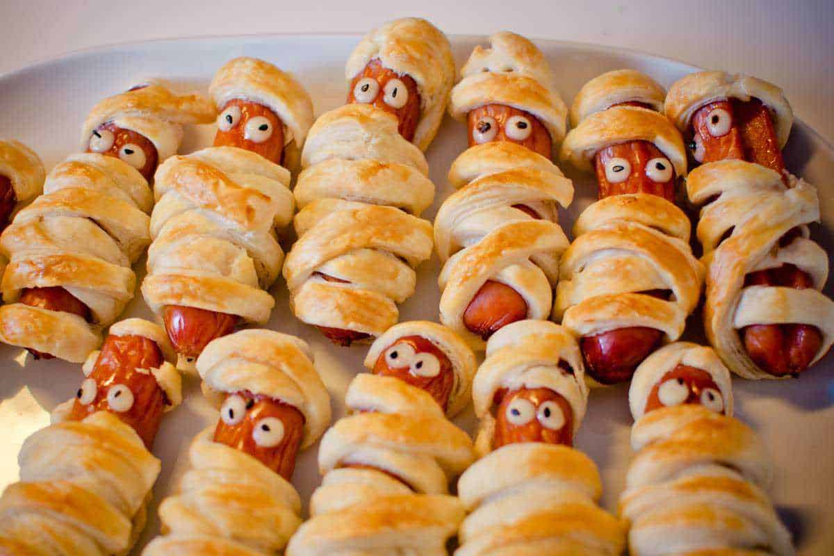 Bastoncini di salsiccia avvolti in pasta sfoglia con occhi di zucchero, preparati come finger food Halloween, presentati su un piatto bianco per una festa spettrale.