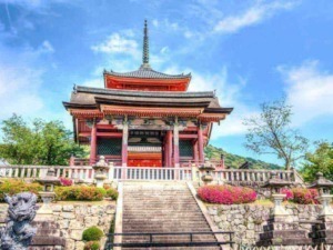 Padiglione tradizionale giapponese in stile pagoda, immerso in un ampio giardino con fiori colorati e alberi, con un cielo azzurro e nuvole leggere sullo sfondo.