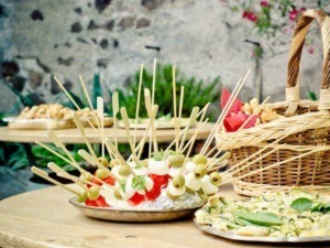Finger food con spiedini di mozzarella, prosciutto e olive, perfetti per aperitivi estivi, con un'ambientazione rustica e naturale.