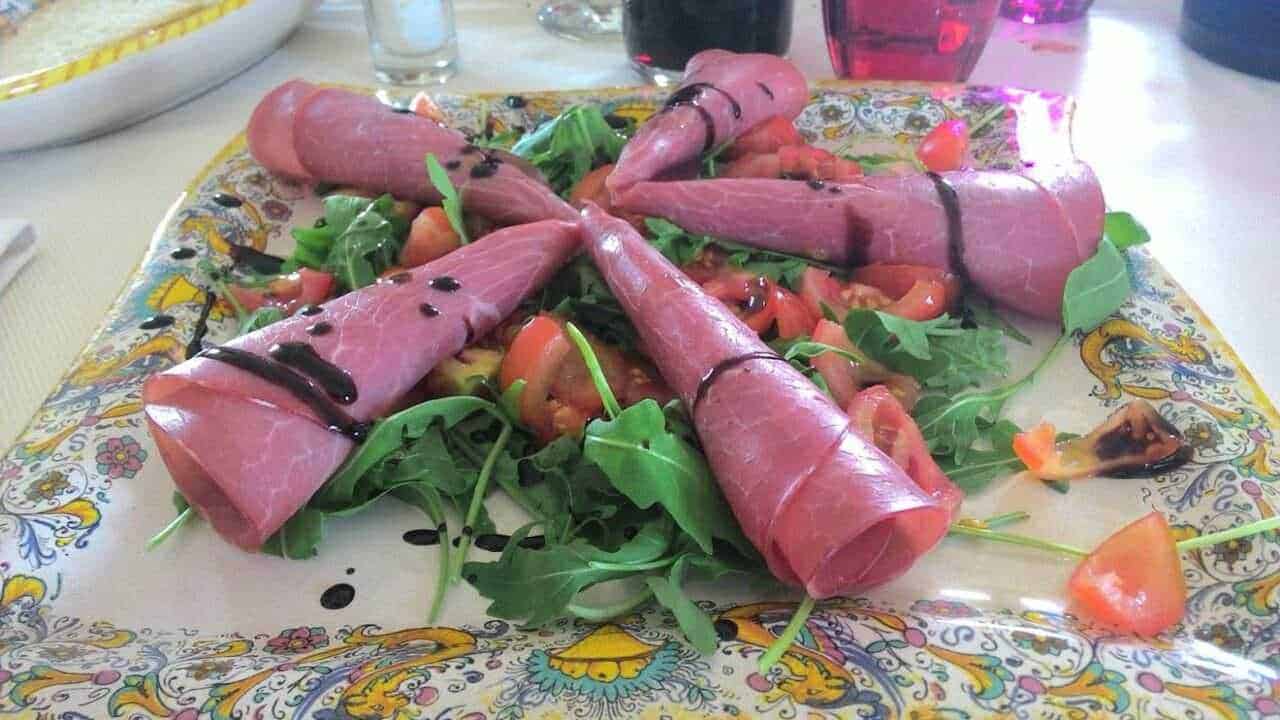 Salumi di prosciutto crudo avvolti intorno a insalata mista con pomodori e salsa balsamica in un piatto decorato tradizionale italiano Altezza, salumi prosciutto crudo, insalata mista, pomodori, salsa balsamica, piatto decorato italiano.