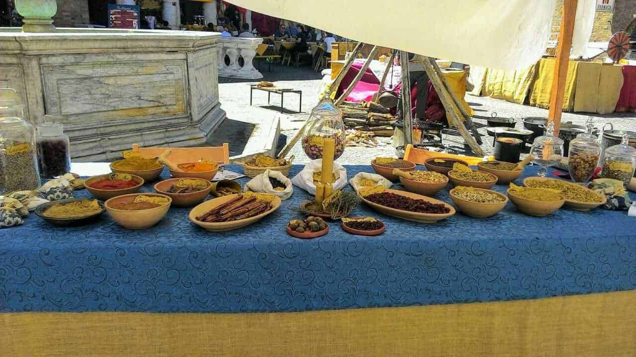 Pane di spezie e dolci tradizionali in esposizione a uno stand di mercato all'aperto. Vari dolci secchi, spezie e confetture presentati in ciotole di ceramica su un tavolo coperto da una tovaglia blu. Ambiente vivace, perfetto per appassionati di gastronomia locale e prodotti tipici.