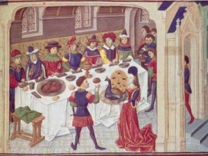 Contenuto dell'immagine di un banchetto medievale con persone in abiti storici, tavolo abbondante di cibo e decorazioni storiche.
