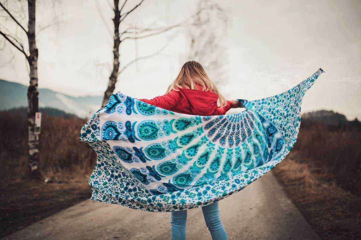 Colorata sciarpa con motivi paisley blu e verde, indossata da una donna in una campagna autunnale, creando un effetto di movimento e libertà, ideale per immagini di moda, natura e stile di vita.