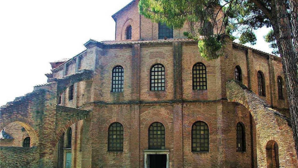 Antico edificio in stile romanico con pareti in mattoni rossi e finestre ad arco, circondato da pini e natura, esempio di architettura storica italiana.