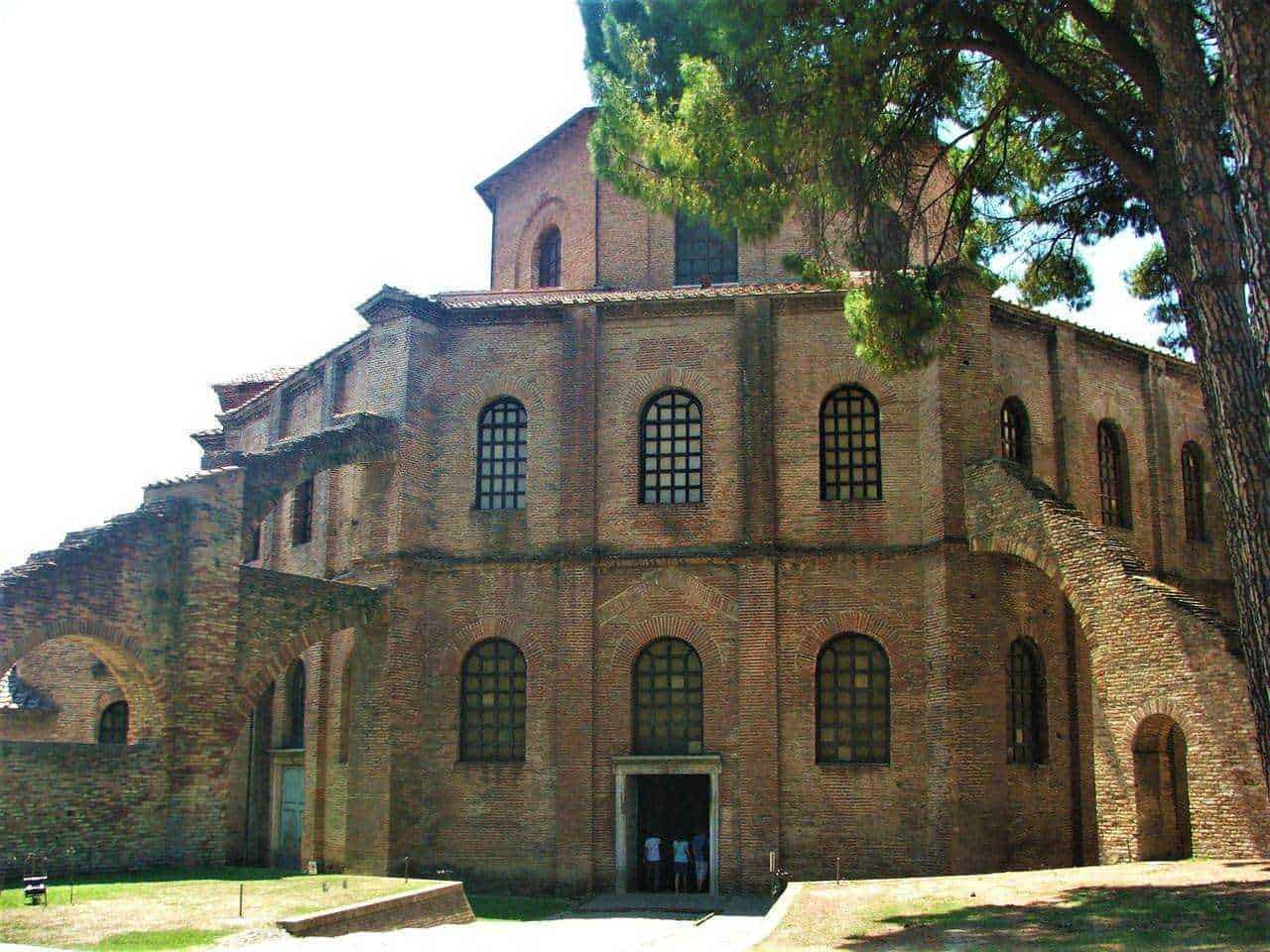 Basilica di san vitale a ravenna: Antico edificio in stile romanico con mattoni a vista e finestre ad arco, circondato da alberi e paesaggio verde.