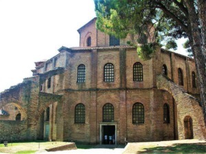 Antico edificio in stile romanico con pareti in mattoni rossi e finestre ad arco, circondato da pini e natura, esempio di architettura storica italiana.