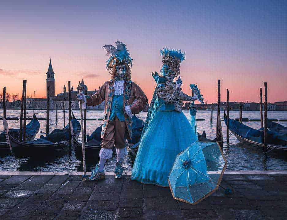 Maschere veneziane tradizionali con costumi eleganti e parasole lungo il canale a Venezia al tramonto. Scene tipiche del Carnevale di Venezia con gondole e skyline storico in sottofondo.