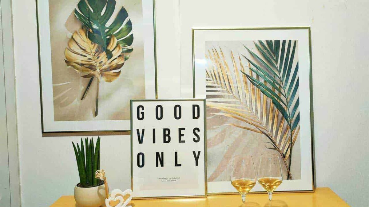 Fogli di monstera e palme in quadri decorativi con frasi motivazionali, pianta grassa e due calici di vino bianco su tavolo in stile boho chic, atmosfera rilassante e trendy per decorazioni d'interni.